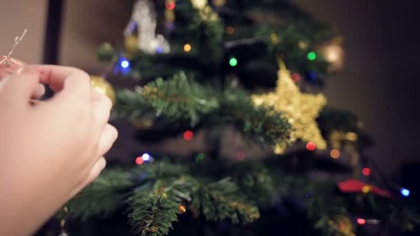 Närbild av en liten flicka dekorerar ett nyårsträd. Hängde leksaker på grenar mot bakgrund av girlanger och ljus — Stockvideo