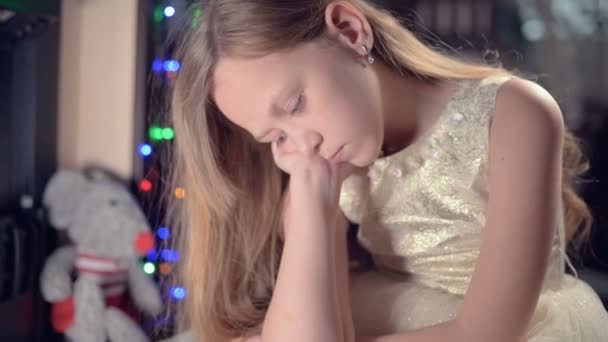 Ein etwas traurig enttäuschtes Mädchen sitzt an einem künstlichen Kamin und ist traurig neben Plüschtieren. das Konzept des verwöhnten Weihnachtsurlaubs — Stockvideo