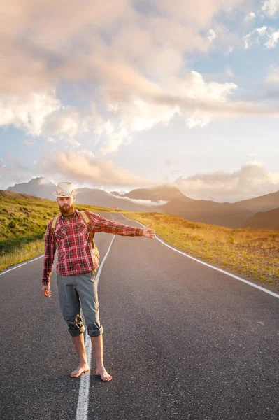 Stilvoller barfüßiger männlicher Tramper mit Hut und Rucksack spaziert bei Sonnenuntergang über eine Asphaltstraße in den Bergen und stoppt das Auto per Anhalter — Stockfoto