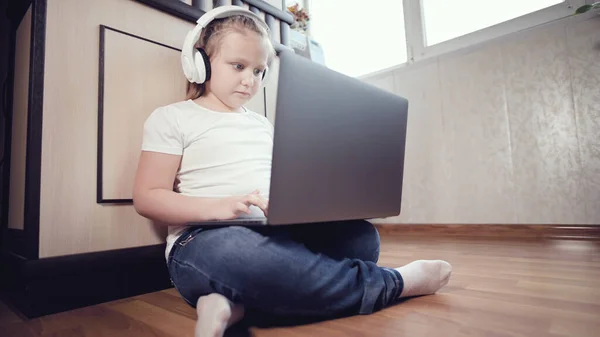 Ένα έξυπνο κοριτσάκι με άσπρα ακουστικά με ένα λάπτοπ στα χέρια σπρώχνει στο πάτωμα στο δωμάτιό της. Η νέα γενιά στο Διαδίκτυο και την τεχνολογία της πληροφορικής — Φωτογραφία Αρχείου