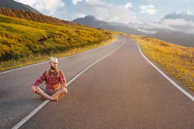 Şık yalınayak sakallı erkek otostopçu bir şapka ve bir sırt çantası ile gün batımında dağlarda bir banliyö asfalt yolda oturur ve gökyüzüne bakarak nakit yardım bekler.