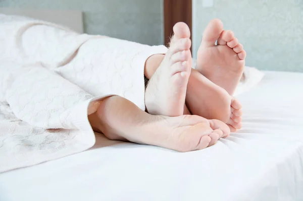 Close-up of the feet of a young couple sticking out from under the covers in the bedroom. Bare feet caress each other engaged in grooming. — Stock Photo, Image