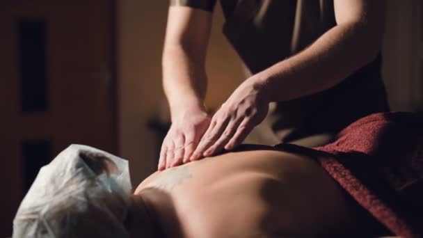 Молодой массажист-мужчина делает массаж спины женщине с татуировкой в массажной комнате с тусклыми лампами на фоне свечей. Низкий ключ премиум массаж — стоковое видео