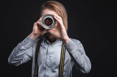 Pantolon askılı, uzun saçlı, sakallı bir adamın stüdyo portresi. Araba parçaları tutuluyor