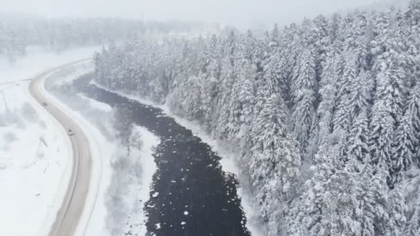 雪の中、川や針葉樹林の近くの冬の国の道路の空中ビュー。冬の天気と冬の降水量の概念 — ストック動画