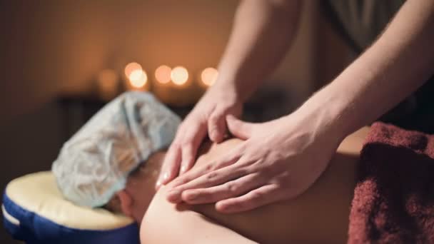 Young male massage therapist does shoulder massage to a woman with a tattoo in a massage room with dim light on the background of candles. Low key concept of premium massage close-up shot — 비디오