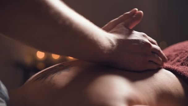 Gros plan Un jeune massothérapeute fait un massage du dos à une femme tatouée dans une salle de massage avec des lumières tamisées sur le fond des bougies. Concept de massage premium low key — Video