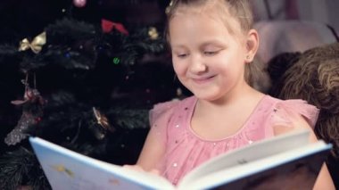 Elinde kitap olan neşeli elbiseli küçük sarışın bir kız Noel ağacının arka planına yaslanmış yumuşak oyuncakların yanına oturur ve kendi evi ile birlikte sayfayı açan bir kitap okur..