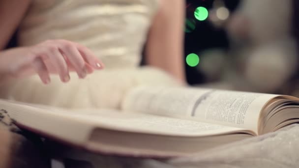 Close-up Una niña rubia en un vestido festivo con un libro en las manos se sienta junto a los juguetes blandos sobre el fondo de un árbol de Navidad y lee un libro que lleva la página con su lugar . — Vídeo de stock