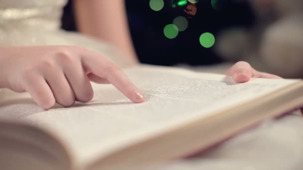 Close-up Uma menina loira em um vestido festivo com um livro em suas mãos se senta ao lado de brinquedos macios contra o fundo de uma árvore de Natal e lê um livro liderando a página com seu lugar . — Vídeo de Stock