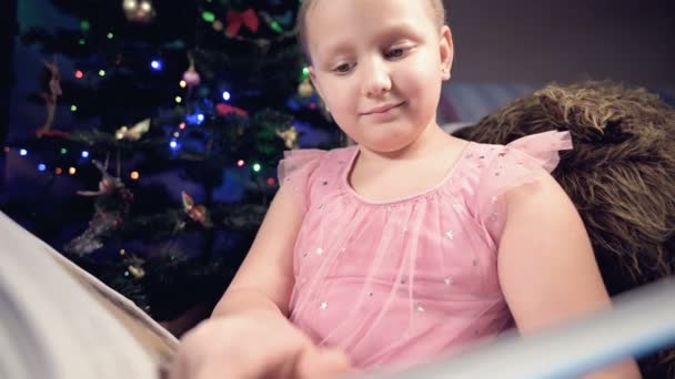 축제 의상을 입고 손에 책을 들고 있는 한 금발 소녀가 크리스마스 트리의 배경에 있는 부드러운 장난감 옆에 앉아서 그녀의 자리와 함께 페이지를 이끄는 책을 읽고 있습니다.. — 비디오
