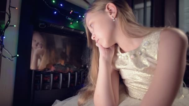 Un po 'triste ragazza delusa siede vicino a un camino artificiale ed è triste accanto a giocattoli morbidi. Il concetto di vacanza di Natale viziato — Video Stock