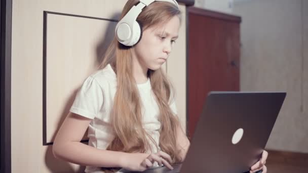 Uma menina esperta de sete anos de idade em fones de ouvido brancos com um laptop em suas mãos está empurrando no chão em seu quarto. A geração jovem na Internet e tecnologia de TI — Vídeo de Stock