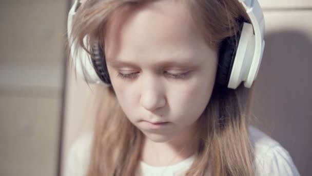 Zbliżenie ujęcia portretu popędliwego planu poważnej zamyślonej i oderwanej dziewczyny, która ma 10 lat w dużych białych słuchawkach w pomieszczeniu. Wygląd boczny — Wideo stockowe