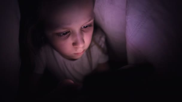 Ένα νεαρό κοριτσάκι κρύβεται κάτω από μια κουβέρτα για να χρησιμοποιήσει μια ψηφιακή συσκευή smartphone tablet αργά μετά τον ύπνο. Μοναξιά των μικρών παιδιών και διάσωση στο τηλέφωνο. — Αρχείο Βίντεο