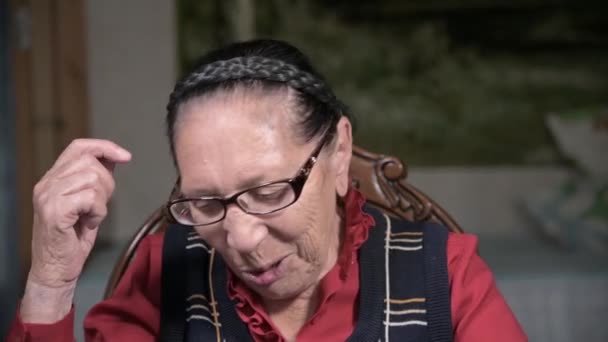 Porträtt av en äldre leende kvinna i glasögon som lutar sig mot armen inomhus och sitter och tänker. Äldre kvinna 80 år — Stockvideo