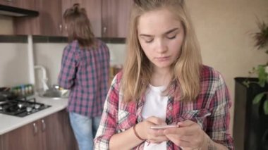 Young married couple in the kitchen. A girl with a tattoo writes a message on her mobile phone to her lover on the background of her husband who washes the dishes.