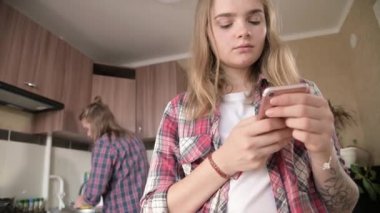 Young married couple in the kitchen. A girl with a tattoo writes a message on her mobile phone to her lover on the background of her husband who washes the dishes.