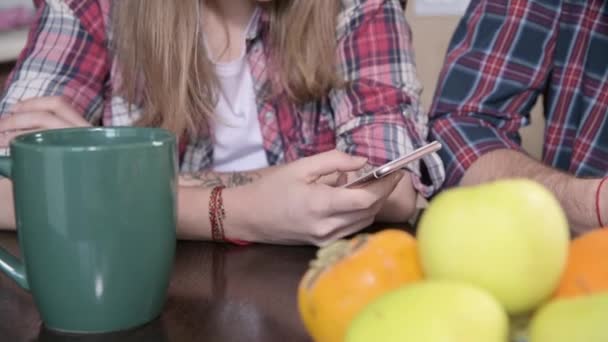 Close-up Un joven par de mellenials con el pelo largo están sentados en la mesa de la cocina con los teléfonos en las manos. Navegación y compras en Internet desde dispositivos móviles. Concepto de familia joven moderna — Vídeos de Stock