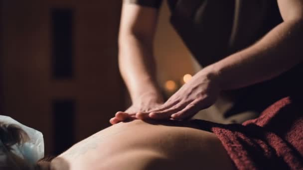 Ung manlig massage terapeut gör rygg massage till en kvinna med en tatuering i ett massagerum med svagt ljus på bakgrunden av ljus. Låg nyckel premium massage koncept — Stockvideo