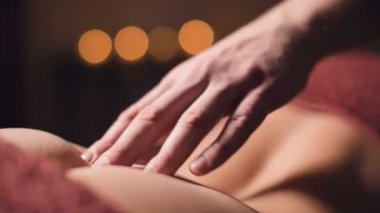 Profesyonel bir fizyoterapistin erkek eli masaj odasında loş ışıkla kadın bir hastaya bel masajı yapıyor.