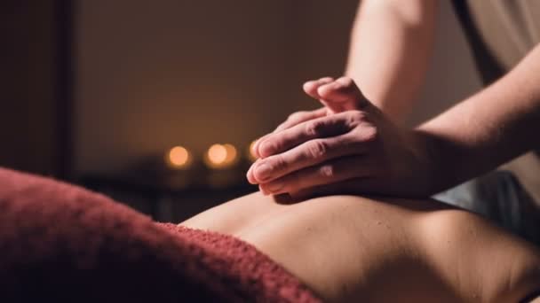 Close-up Jovem massagista do sexo masculino faz massagem nas costas para uma mulher com uma tatuagem em uma sala de massagem com luzes escuras no fundo das velas. Conceito de massagem premium de tecla baixa — Vídeo de Stock