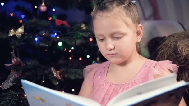 Ένα μικρό ξανθό κορίτσι σε ένα εορταστικό φόρεμα με ένα βιβλίο στα χέρια της κάθεται δίπλα σε μαλακά παιχνίδια στο φόντο ενός χριστουγεννιάτικου δέντρου και διαβάζει ένα βιβλίο που οδηγεί τη σελίδα με τη θέση της. — Αρχείο Βίντεο