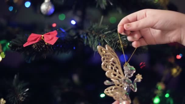 Крупный план руки маленькой девочки украшает новогоднюю елку. Повешивает игрушки на ветках на фоне гирлянд и огней — стоковое видео