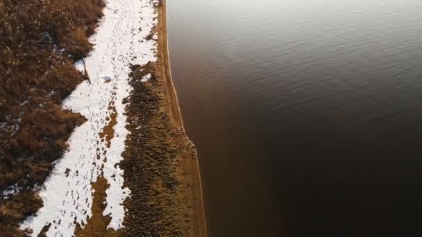 Vue aérienne du haut du rivage par une chaude journée d'hiver. Herbe jaune et morceaux de neige sur le rivage. Concept de réchauffement climatique. Mouvement en avant — Video