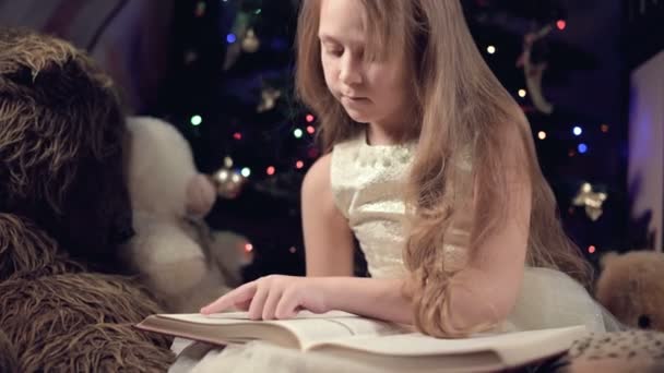 Una niña rubia en un vestido festivo con un libro en las manos se sienta junto a juguetes suaves sobre el fondo de un árbol de Navidad y lee un libro que lleva la página con su lugar. — Vídeos de Stock