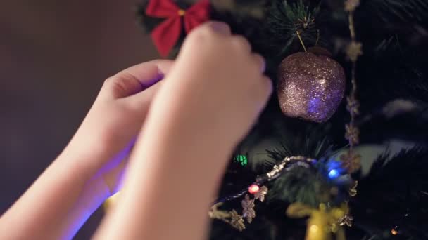 Крупный план руки маленькой девочки украшает новогоднюю елку. Повешивает игрушки на ветках на фоне гирлянд и огней — стоковое видео