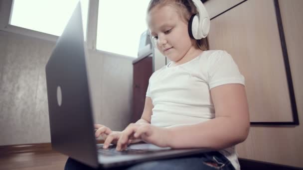 Una niña inteligente de siete años con auriculares blancos con un portátil en las manos está empujando al suelo en su habitación. La generación joven en Internet y la tecnología informática — Vídeo de stock