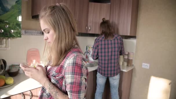 Casamento jovem na cozinha. Uma menina com uma tatuagem escreve uma mensagem em seu telefone celular para seu amante no fundo de seu marido que lava a louça . — Vídeo de Stock