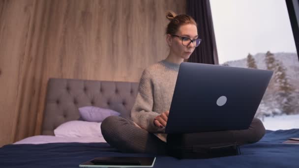 Portrait d'une jolie femme indépendante avec des lunettes et un pull avec des bas assis sur un lit dans une éco-maison au milieu d'une forêt d'hiver avec un ordinateur portable — Video