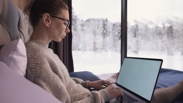 Portrait of attractive freelancer woman with glasses and a sweater with stockings sitting on a bed in an eco-house in the middle of a winter forest with a laptop — Stock Video