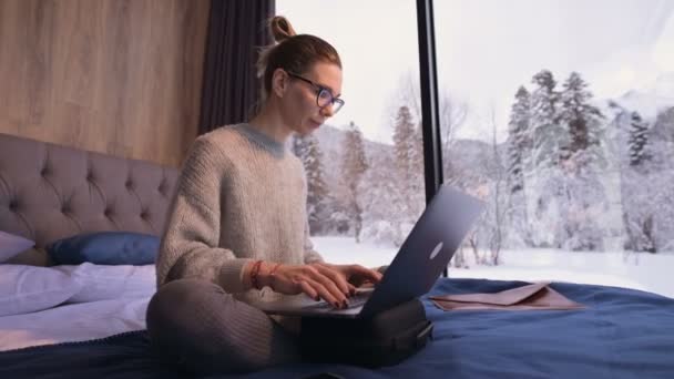 Retrato de mulher freelancer atraente com óculos e uma camisola com meias sentadas em uma cama em uma eco-casa no meio de uma floresta de inverno com um laptop — Vídeo de Stock