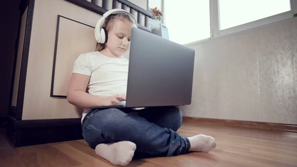 Elinde dizüstü bilgisayarla beyaz kulaklıklı akıllı küçük bir kız odasında yere bastırıyor. İnternet ve bilişim teknolojisindeki genç nesil. — Stok fotoğraf