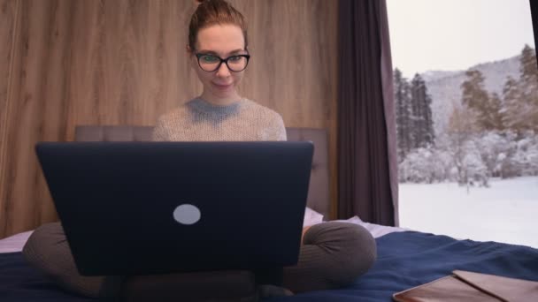 Retrato de una atractiva mujer freelancer con gafas y un suéter con medias sentado en una cama en una eco-casa en medio de un bosque de invierno con un ordenador portátil — Vídeo de stock