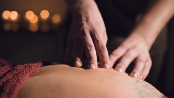Junge männliche Massagetherapeut macht Fingermassage einer Frau mit einer Tätowierung in einem Massageraum mit schummrigem Licht auf dem Hintergrund von Kerzen. Günstiges Premium-Massagekonzept — Stockvideo
