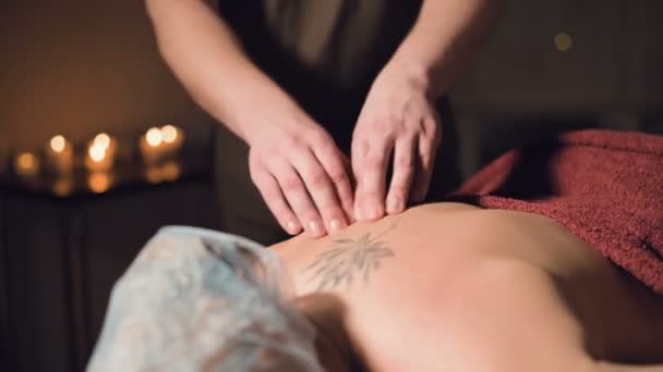 Jonge mannelijke massage therapeut doet vinger massage van een vrouw terug met een tatoeage in een massage kamer met schemerig licht op de achtergrond van kaarsen. Low-key premium massageconcept — Stockvideo