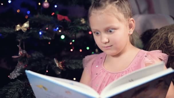 Een klein blond meisje in een feestelijke jurk met een boek in haar handen zit naast zacht speelgoed tegen de achtergrond van een kerstboom en leest een boek leidt de pagina met haar plaats. — Stockvideo