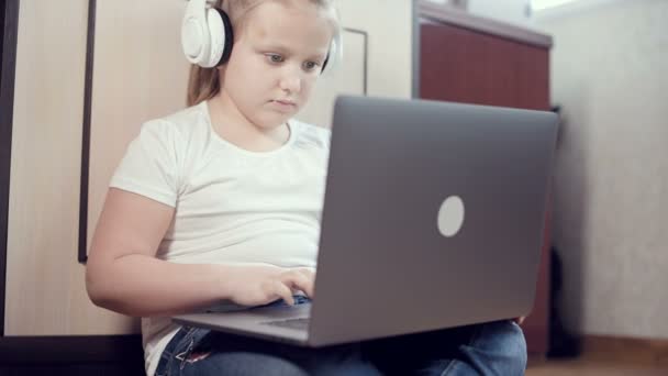 Uma menina esperta de sete anos de idade em fones de ouvido brancos com um laptop em suas mãos está empurrando no chão em seu quarto. A geração jovem na Internet e tecnologia de TI — Vídeo de Stock