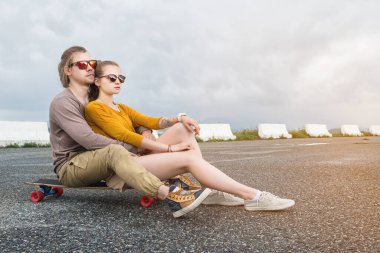 Güneş gözlüklü çekici, genç, uzun saçlı bir çift banliyö asfalt yolundaki kaykayın üzerinde güzel bulutlu bir gökyüzüne sarılıyor..