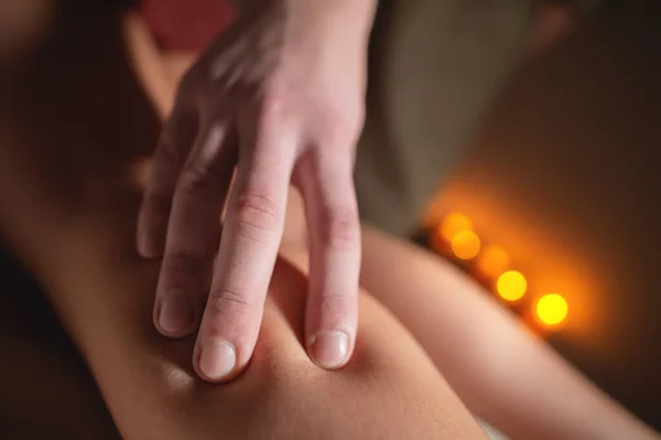 Close-up hand of a massage therapist massaging the legs of a female client against the background of burning candles in an office with a cozy light. — Stock Photo, Image