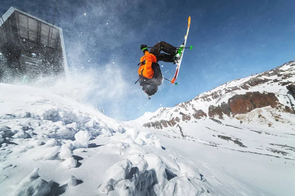 Esquiador de visão de ângulo baixo atleta em uma jaqueta laranja faz uma volta com pó voador de neve contra um céu azul claro e montanhas cobertas de neve do Cáucaso . — Fotografia de Stock