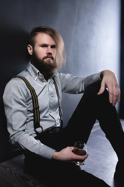 Retrato del estudio de un hombre con estilo barbudo con un corte de pelo de moda en una camisa y tirantes con una copa de vino o coñac en la mano. — Foto de Stock