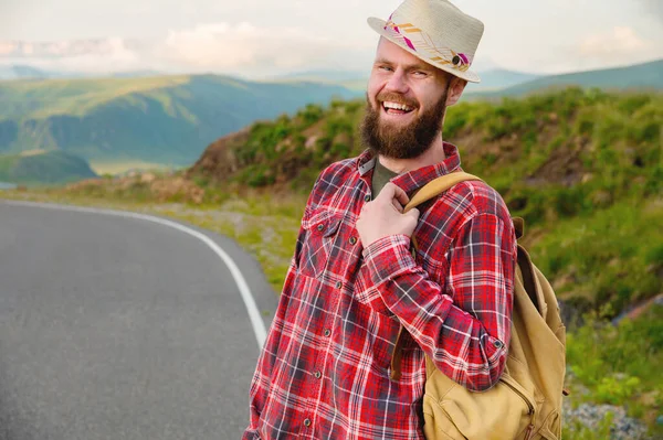 Retrato de um alegre sorridente barbudo viajante homem em um chapéu e uma camisa xadrez com uma mochila em seu ombro na natureza contra o fundo de campos verdes do céu e nuvens . — Fotografia de Stock