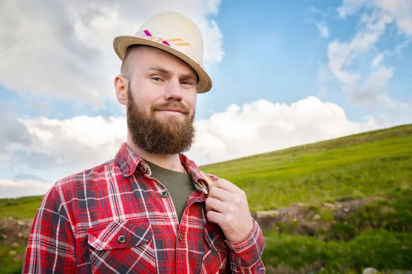 Retrato de un alegre viajero barbudo sonriente con un sombrero y una camisa a cuadros con una mochila en su hombro sobre la naturaleza sobre el fondo de campos verdes del cielo y las nubes . — Foto de Stock