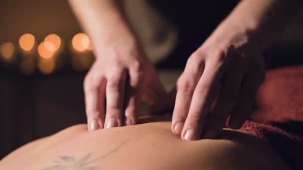 Tânărul terapeut de masaj masculin face masaj cu degetul al unei femei înapoi cu un tatuaj într-o cameră de masaj cu lumină slabă pe fundalul lumânărilor. Conceptul de masaj premium cu cheie redusă — Videoclip de stoc