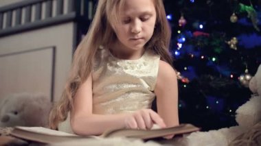 Elinde kitap olan neşeli elbiseli küçük sarışın bir kız Noel ağacının arka planına yaslanmış yumuşak oyuncakların yanına oturur ve kendi evi ile birlikte sayfayı açan bir kitap okur..